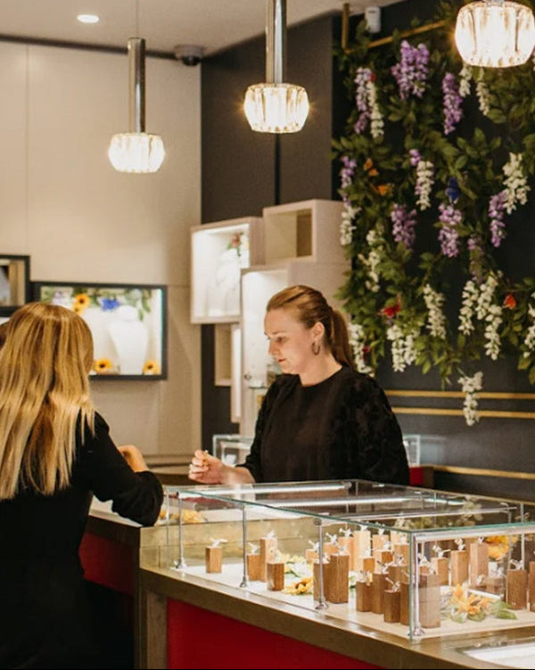 Two women in a store with jewelry display cases and floral decorations.